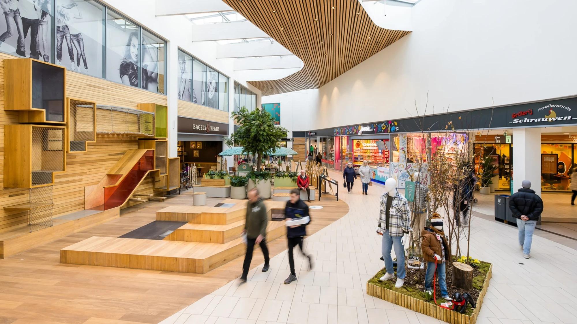 Overdekt winkelcentrum met houten zitelementen, groen en winkelend publiek